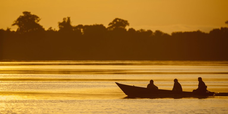 rio margeado pela floresta ao entardecer. Sobre o rio, do lado esquerdo, está uma rabeta com três pessoas sentadas.