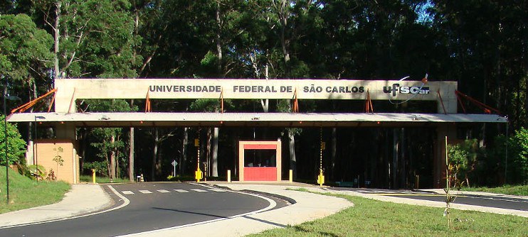 visão frontal do portal da entrada principal da Universidade Federal de São Carlos, composto pelo nome da universidade, uma guarita e árvores ao fundo.